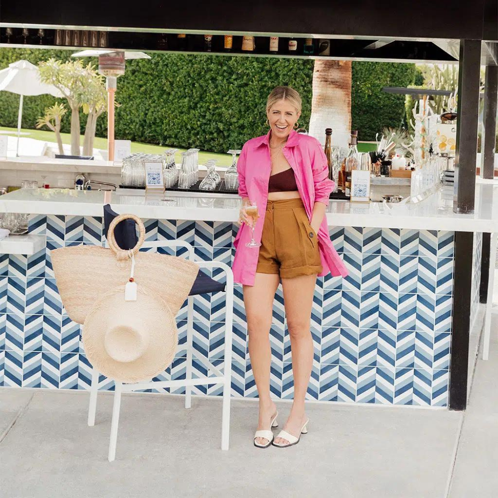 Woman standing by bar in pink jacket, holding a drink, next to a chair with a tote bag and hat clipped by a TOPTOTE hat holder
