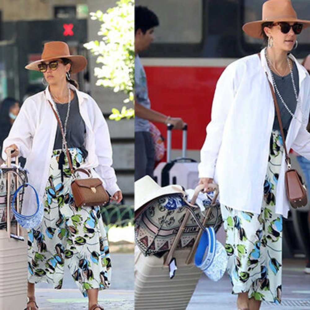 Celebrity spotted in Florence, Italy, wearing a floral skirt, white jacket, and brown hat, pulling luggage on a sunny summer day
