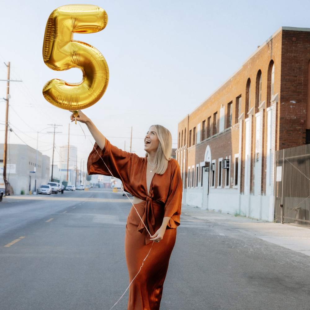 Woman celebrating 5 years in business holding a gold number five balloon in an urban setting.