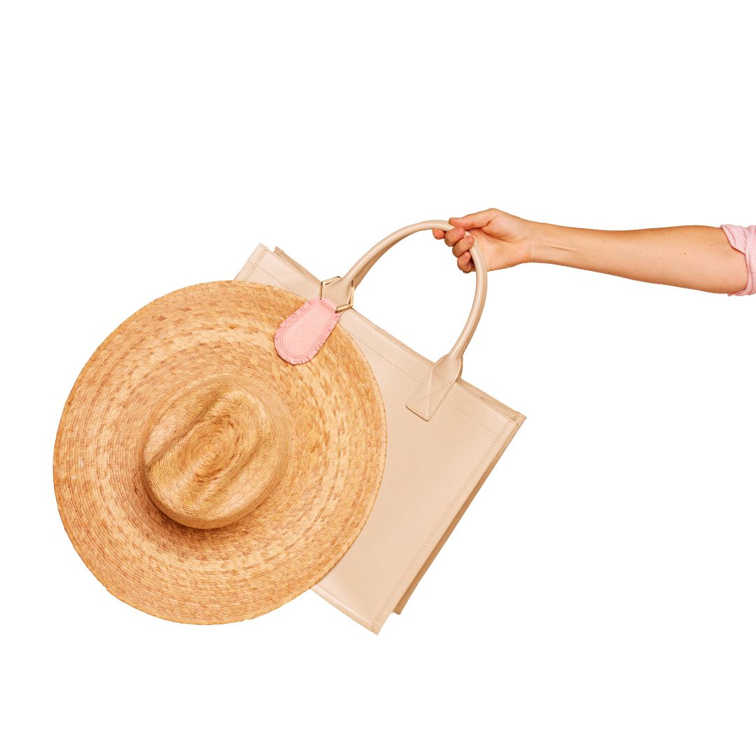 Person holding a beige bag with a straw hat attached using The Fray TOPTOTE Hat Clip with raffia trim detailing.