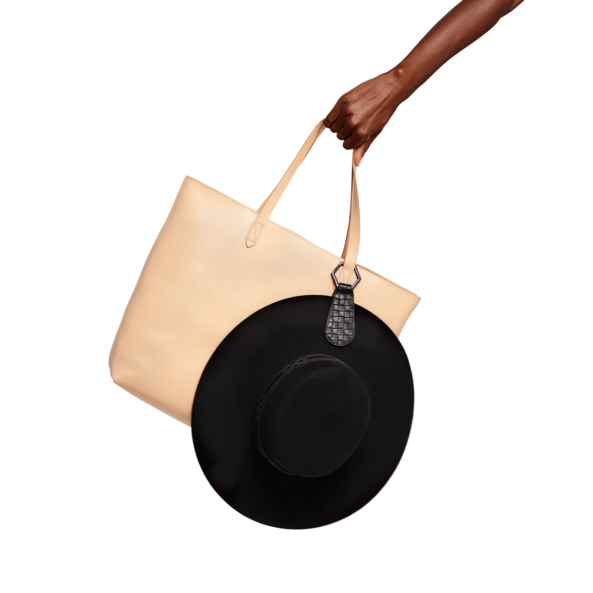 Person holding beige tote bag with black hat attached using The Drop TOPTOTE hat clip for hands-free travel.