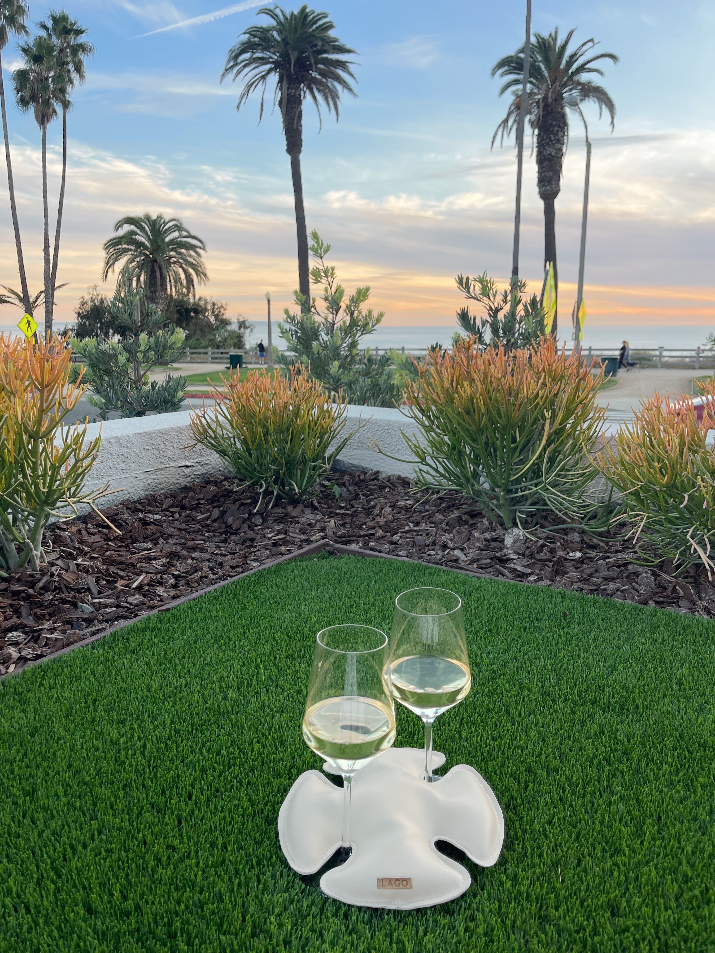 Wine glasses securely held by Lago 'Sand Dollar' Wine Holder on grass, with sunset and palm trees in the background.