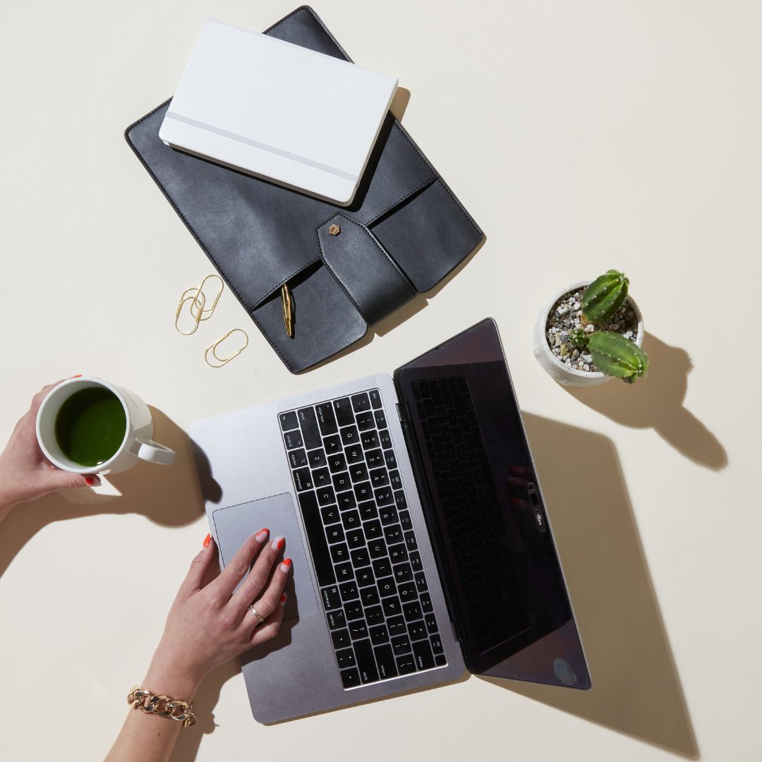 Person working on a laptop with a notebook, leather portfolio, paper clips, coffee cup, and small potted cactus on a white desk