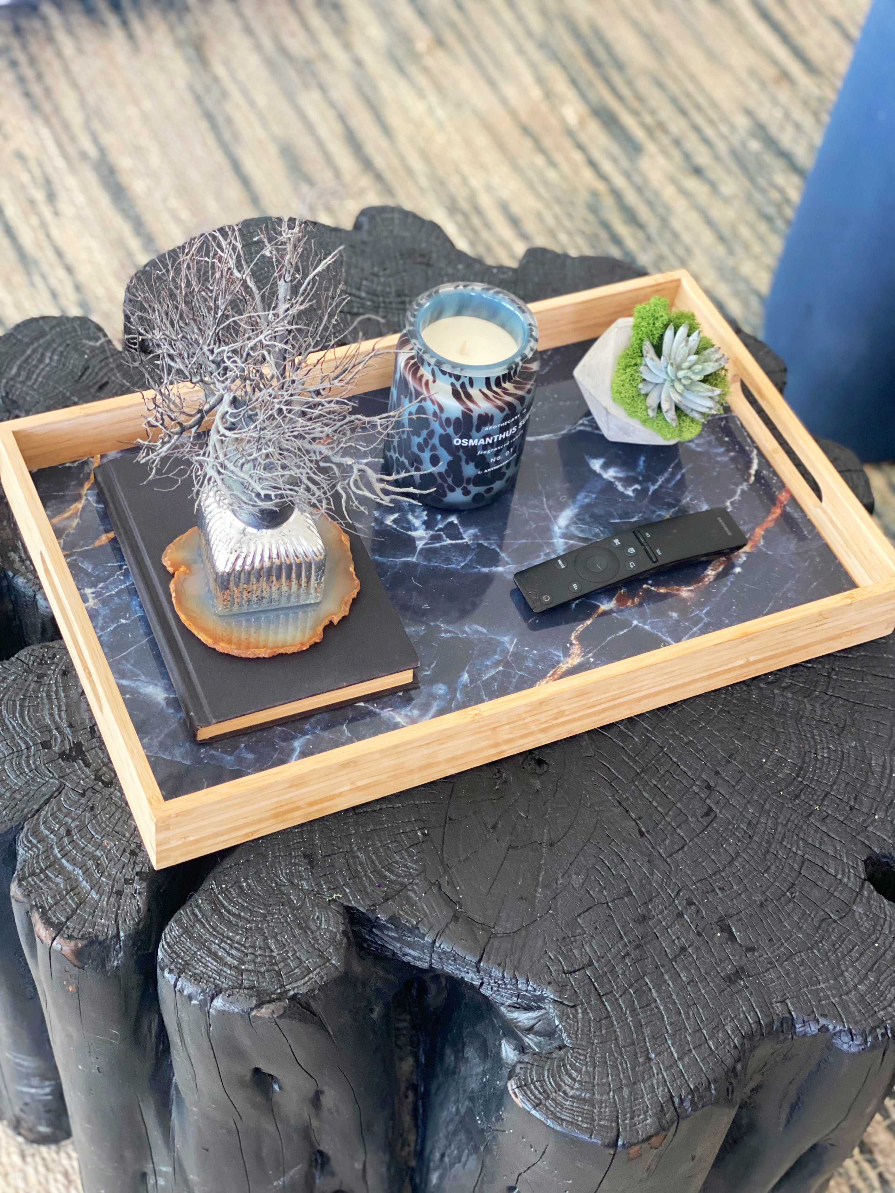 Wooden tray displaying candle, remote, plant, and decor on black marble insert, atop textured dark table.