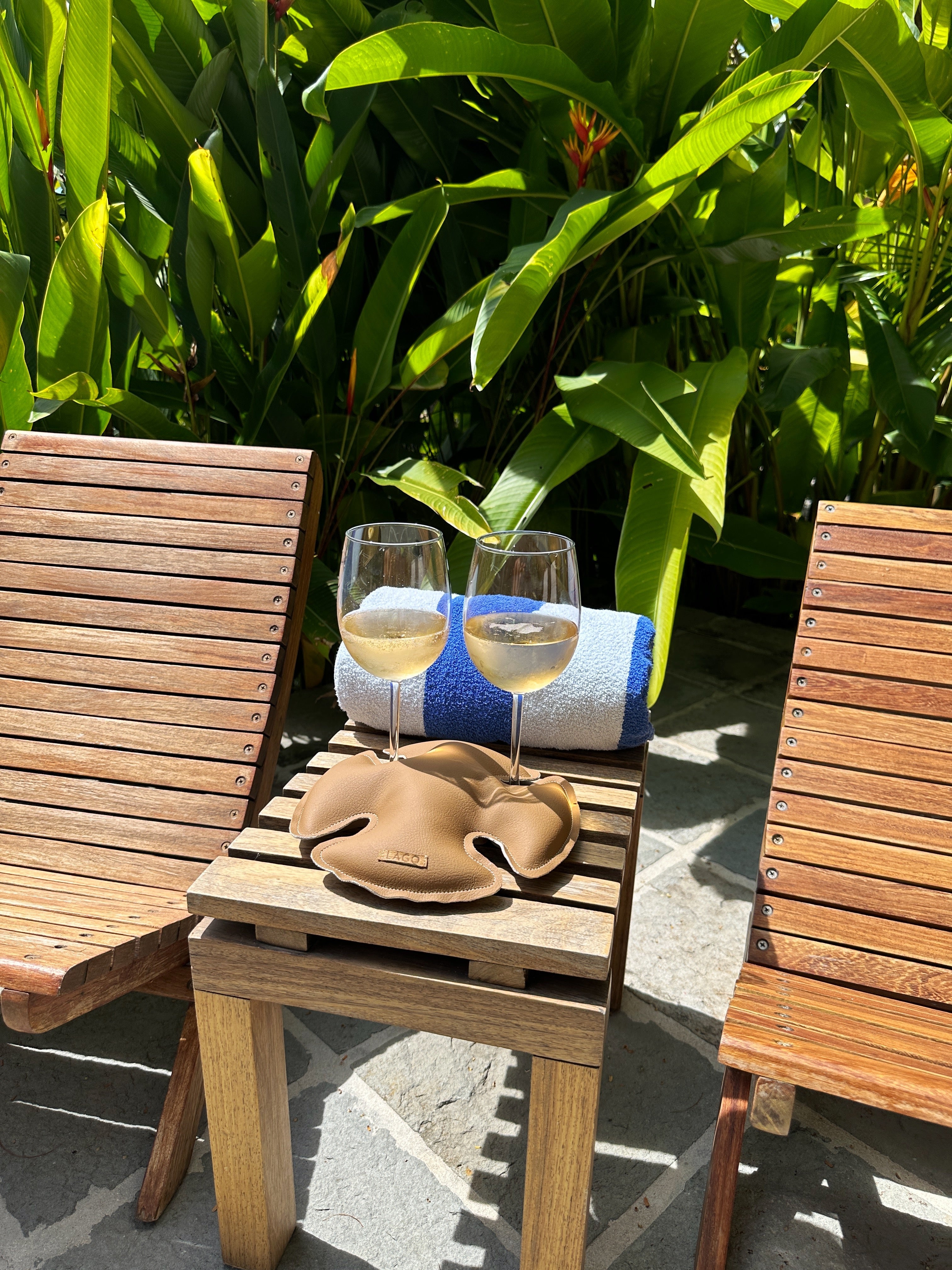 Two glasses of chilled wine poised on a LAGO stabilizer between wooden chairs, ready for a relaxing poolside retreat amidst lush tropical foliage.