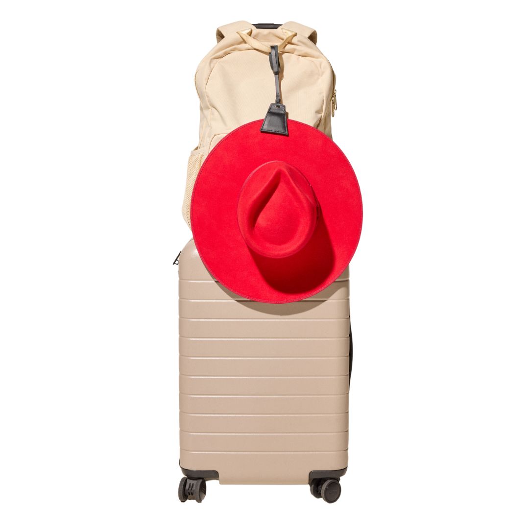 Lasso TOPTOTE hat clip in black holding a red hat and attached to a cream backpack that is resting on a beige carry-on suitcase.